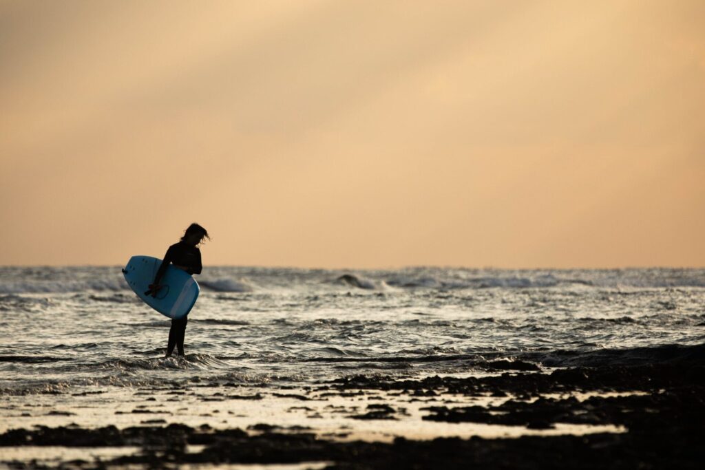 夕方の海辺でサーフィンボートを持ちながら歩くサーファー